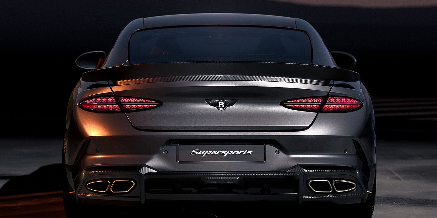 Bach Premium Cars GmbH | Bentley Mannheim Bentley Supersports rear view with illuminated rear lamps, spoiler and black Bentley badge in Anthracite paint against a dark grey and red background