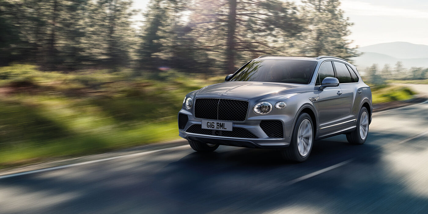 Bach Premium Cars GmbH | Bentley Mannheim Bentley Bentayga luxury SUV front three quarter in Moonbeam paint, driving on an country road past trees.